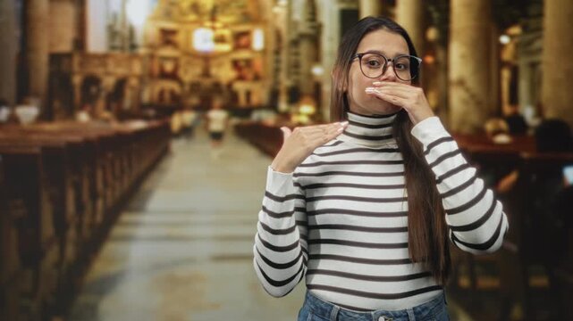 Woman covering mouth with hand and pointing to throat while wearing glasses and striped turtleneck inside a building with pews and altar; silence reverence.