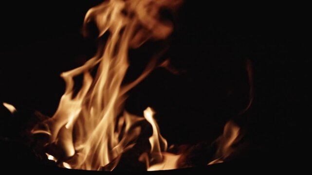 Cinematic slow-motion footage of a flickering fire in a backyard fire pit, featuring detailed close-ups of glowing orange embers and wide shots of dancing flames against a dark evening background.