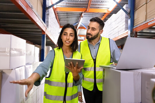 Two logistics workers in uniforms standing in warehouse aisle, discussing inventory, reviewing stock data, or checking products quantity. Teamwork, coordination of supply chain, stocktaking management
