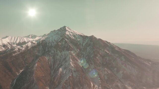 Drone footage of snow-dusted Peak Galina rising above Arshan village in Buryatia, Russia, with bright sun and lens flare over rugged ridgelines and valleys