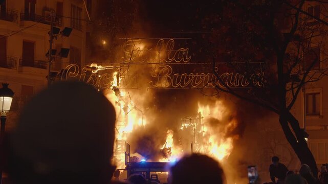 Large falla sculpture consumed by intense orange flames during Las Fallas festival in Valencia, illuminated decorative cursive sign above fire as crowd silhouettes watch from darkness