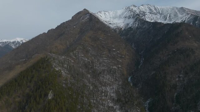 Drone footage of Pik Lyubvi mountain ridge with conifer forest and patchy snow on steep slopes near Arshan, Republic of Buryatia, Russia, under cloudy sky