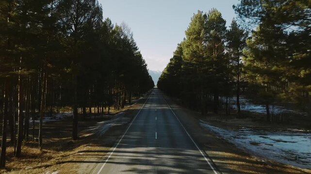 Aerial view of a straight two lane road cutting through dense pine forest with patches of snow, leading toward mountains near Arshan, Buryatia, Russia
