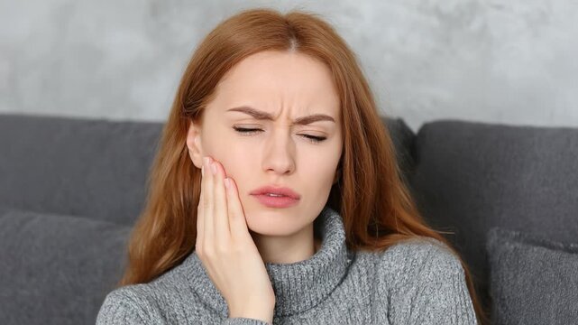 A Young Woman in a Gray Sweater Exhibits Distress Due to Tooth Pain, Gripping Her Jaw in Discomfort While Sitting on a Sofa with a Neutral Background