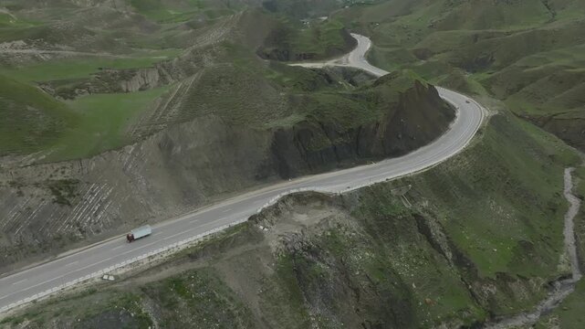 Drone footage of a serpentine asphalt road cutting through green mountain slopes, with a cargo truck driving along the curve in Dagestan, Russia, scenic travel route