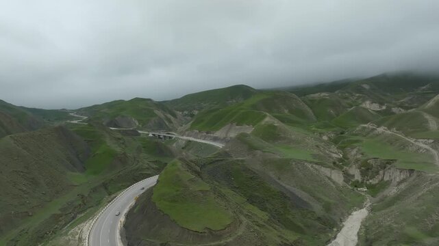 Drone footage of a curving highway with cars crossing a mountain valley among lush green hills under cloudy sky in Dagestan, North Caucasus, Russia