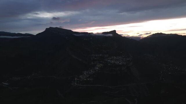 Drone footage of Gunib village on a mountain plateau at sunset, with dramatic clouds, winding roads and valley lights, Republic of Dagestan, Russia