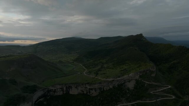 Drone footage of a winding mountain road leading to Keger village across green ridges and cliffs at dusk under dramatic clouds, Dagestan, Russia