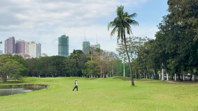Filipino Golf Caddy Walking Across Green Fairway with City Skyline Background