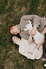 Naklejka premium Natural, smiling couple resting on grass with a playful goat kid, celebrating outdoor lifestyle and rustic charm. Cozy sweaters, gentle light, candid affection, relaxed mood, and connection with
