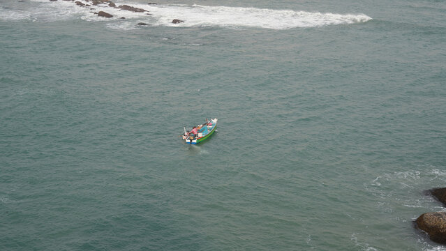 Aerial view of a colorful traditional fishing boat on turquoise sea water in Kanyakumari, Tamil Nadu, India, with distant fishermen working onboard near the coastal shoreline.