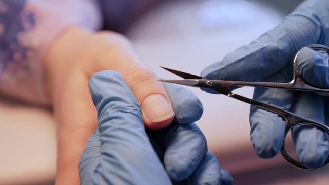 Macro salon close-up of a nail technician trimming the cuticle edge with fine scissors, showing precise dry manicure prep, hygiene, and careful professional nail care.