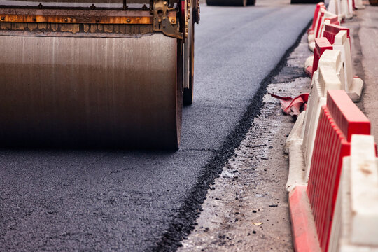 Workers are paving asphalt on a city street, with a roller compactor smoothing out the surface. Road barriers are visible, indicating an active construction zone in an urban setting.