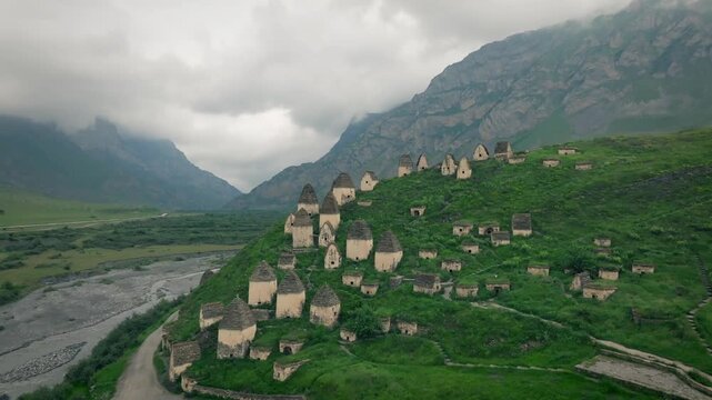 Drone footage of the Dargavs necropolis with ancient stone crypts on a green hillside under cloudy sky, North Ossetia&ndash;Alania, Russia, Caucasus Mountains