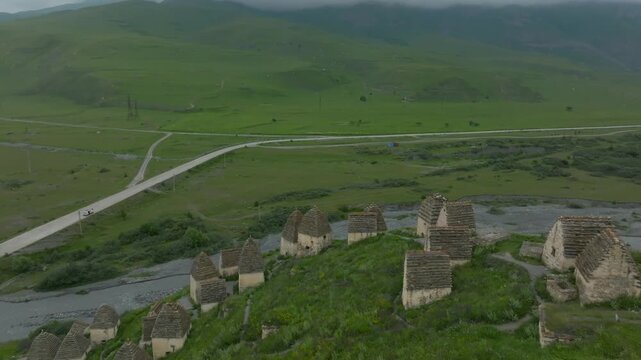 Drone footage of the Dargavs necropolis with ancient stone crypts on a green hillside above a river and road in North Ossetia&ndash;Alania, Russia, cloudy day