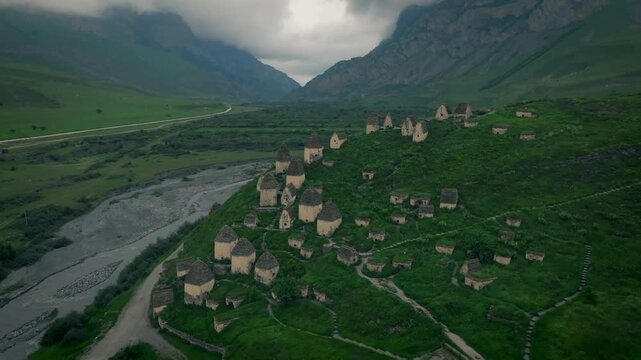 Drone footage of the Dargavs City of the Dead necropolis with ancient stone crypts on green hillside beside a river, North Ossetia&ndash;Alania, Russia