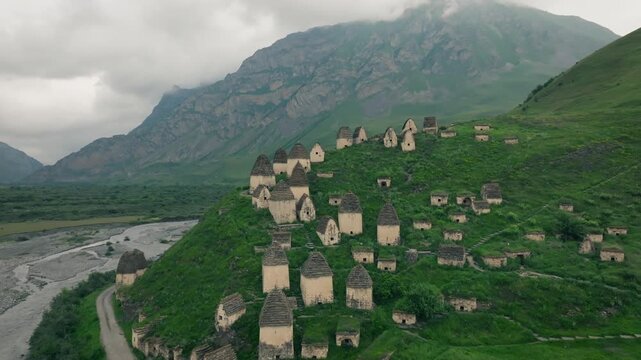 Drone footage of the Dargavs necropolis with ancient stone crypts on a green hillside above a river valley, North Ossetia&ndash;Alania, Russia under cloudy sky