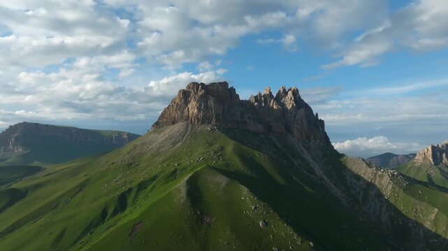 Scenic mountain landscape with jagged rocky summit and lush green slopes under moving clouds, view of Mount Naudzhidza in Russia, wide outdoor nature panorama