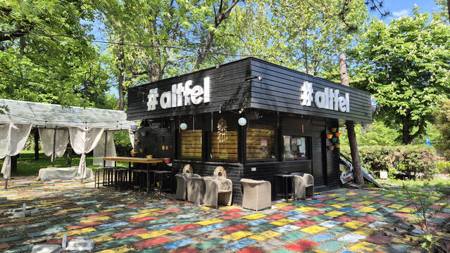 A black wooden kiosk/restaurant named #altfel in Titan park with colorful pavement, and green trees. Picture taken on May 5, 2026 in Bucharest, Romania.