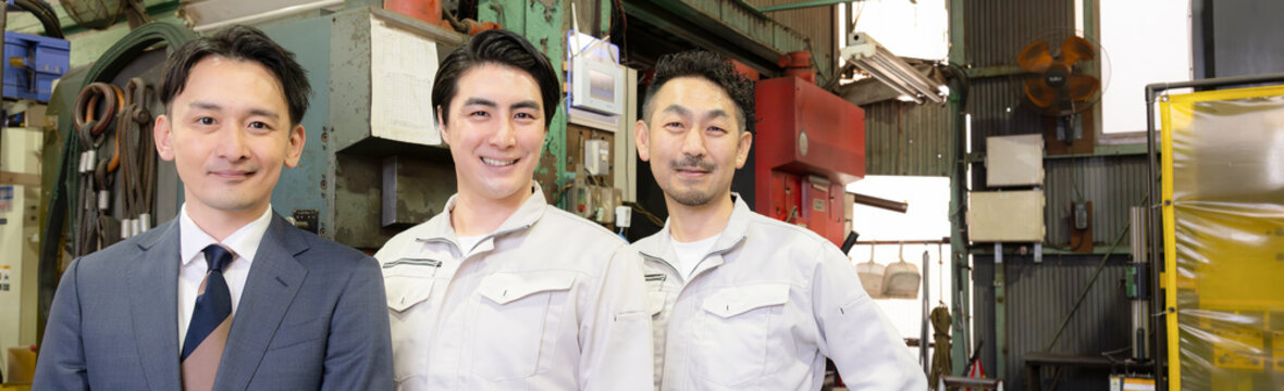 工場の製造業の現場で働く作業着とスーツの営業マン3人チームのポートレート　中小企業のカメラ目線の笑顔のバナー