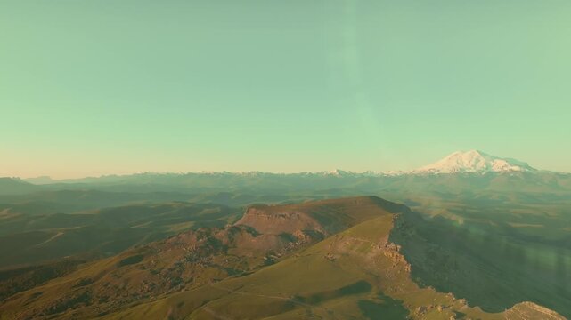 Aerial footage of Bermamyt Plateau with rugged cliffs and green ridges, with snowcapped Mount Elbrus on the horizon in Karachay-Cherkessia, Russia