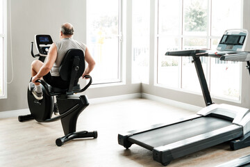 Fototapeta premium Man training on a recumbent exercise bike in a bright home gym with a treadmill and large windows