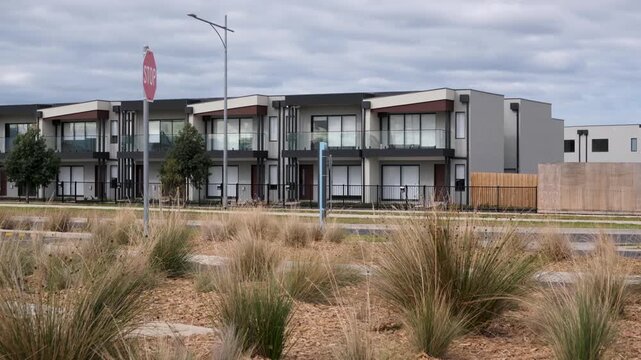 A row of newly constructed, contemporary two-storey townhouses in a modern Australian suburban residential estate, against dry woodchip mulch and clumps of drought-resistant native tussock grasses