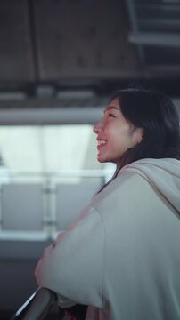 Happy young Asian woman with a hopeful smile looking up while standing on a city skywalk at night. Wearing a beige hoodie, she reflects on life amidst urban bokeh lights. Cinematic