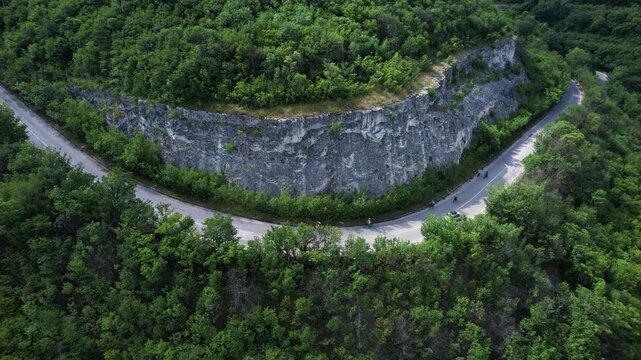 Aerial view of cyclists riding along a winding forest road next to a steep limestone cliff face in Provadia, Varna, Bulgaria.