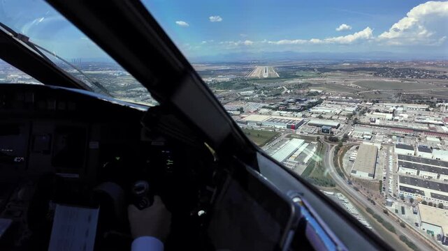 Immersive pilot view through the cockpit windshield of the approach to Madrid airport runway 32R, with a view of copilot hand at the controls, and the runway ahead under a blue sky with few clouds