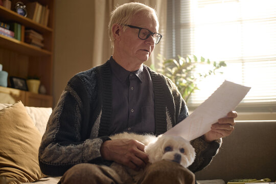 Senior Caucasian man reading document while sitting with pet dog, reviewing household paperwork and finances at home. Useful for retirement planning, insurance, billing, budgeting themes