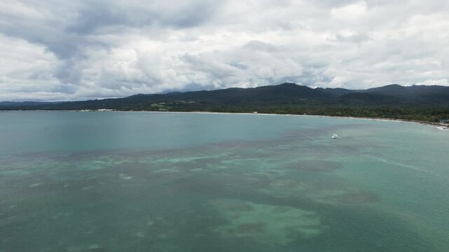 Aerial view of the turquoise coastline and a boat near the shore with lush green hills under a cloudy sky in Wewak, East Sepik Province, Papua New Guinea.