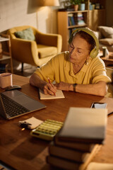 Obraz premium Vertical shot of senior Caucasian woman wearing headphones, writing notes beside laptop and calculator at home. Supporting online learning, remote training, financial planning content
