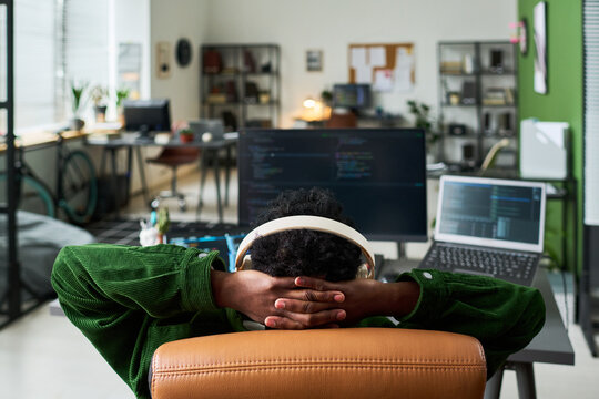 Young African American man reclining at coding workstation, wearing headphones and resting after programming session. Ideal for remote work, developer burnout, tech productivity