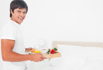 Man standing left in bedroom holding wooden tray with juice, croissant, yogurt, rose, copy space © wavebreak3