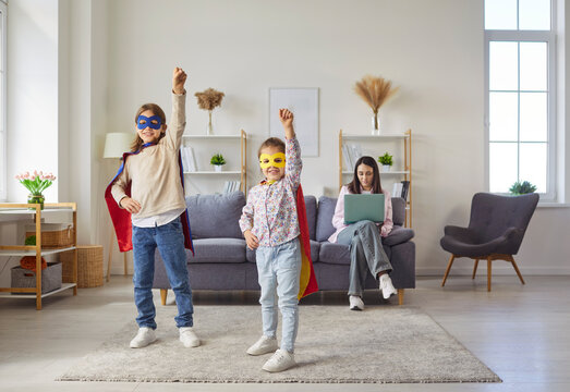 Happy girl and boy in masks and capes of superhero play together in home living room while mother working on laptop. Smiling super kids standing on carpet with hands up, pretend brave strong heroes