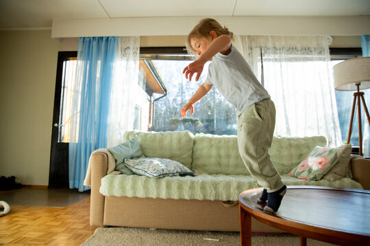 Low angle shot of joyful small child with long hair jumping and soaring off sofa, full of energy and playful laughter during a bright morning at home