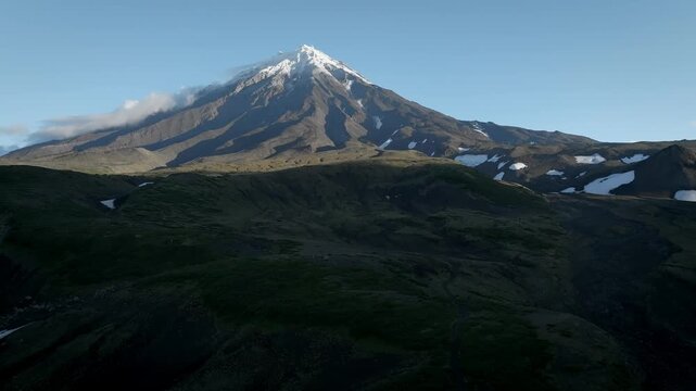 Aerial footage of Koryaksky Volcano with snow on the summit and drifting clouds above rugged green tundra hills, Kamchatka Krai, Russia, on a clear day