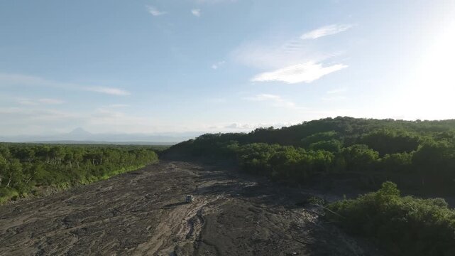 Aerial footage of an off-road vehicle driving along the wide Sukhaya riverbed road toward Avachinsky Volcano, Kamchatka Krai, Russia, with forest and mountains