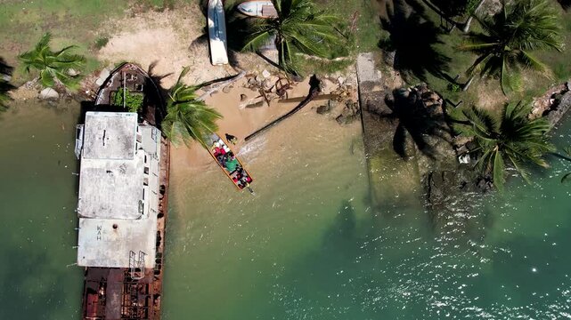 Aerial view of a large rusty shipwreck and a colorful boat with people near a sandy shoreline with palm trees in Wewak, East Sepik Province, Papua New Guinea.