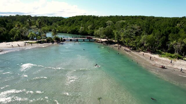Aerial view of people swimming in turquoise coastal waters near a bridge and dense tropical forest with a sandy beach Wewak, East Sepik Province, Papua New Guinea.