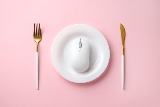 White wireless computer mouse on a white plate, flanked by gold fork and knife on pink background, creative flat lay concept. Top view.