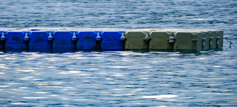 Close up view of a modular floating pontoon dock constructed from interlocking blue and grey plastic cube blocks resting on the gentle ripples of a calm reflective marine environment