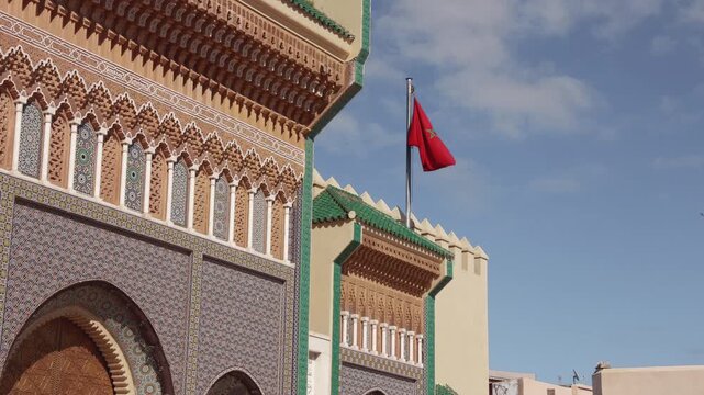 Detail of a traditional Moroccan building with intricate zellige tile work. The red flag of Morocco is waving in the wind under a blue sky