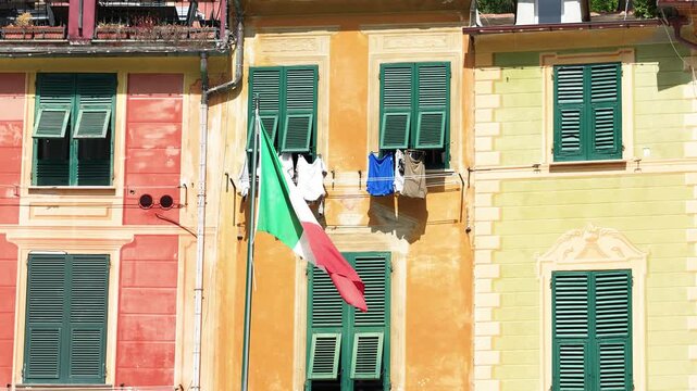 Vivid Italian flag flutters before charming pastel buildings, green shutters, and hanging laundry in bright sunlight. Crisp, steady composition, sharp focus, perfect for stock.