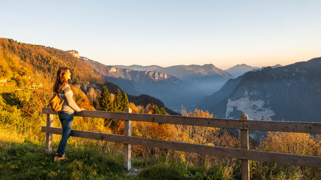 Woman with backpack enjoying a scenic mountain view during golden hour, surrounded by autumn foliage and a valley, promoting travel and exploration in Luserna, Trentino, Italy.
