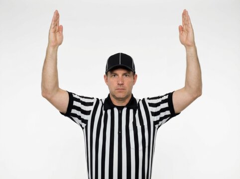 An American football referee, clad in a striped uniform and cap, confidently raises both arms straight upwards to signal a score has been successfully achieved during a competitive sporting