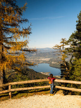 Outdoor travel woman with backpack enjoys panoramic autumn mountain view of lake and town from a viewpoint in Trentino, Italy, representing freedom and adventure.