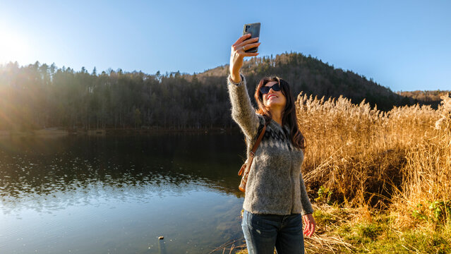 Woman taking a selfie with smartphone by scenic Lavarone lake, enjoying outdoor lifestyle, travel, and relaxation in Paganella, Trentino, Italy.