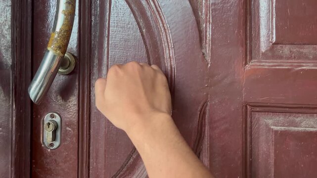 Close up of a fist knocking on a dark brown wooden door with peeling paint handle.